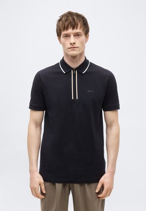 Mladý muž stojaci, oblečený v čiernej krátkorukávovej polo košeli s bielym lemovaným golierom a zipsovou légou, v kombinácii s béžovými nohavicami.