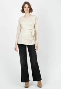 Beige Langarmoberteil mit Wickeldesign und seitlichem Bindeband, kombiniert mit schwarzen Schlagjeans und beigefarbenen Stiefeletten, vor einem schlichten weißen Hintergrund.
