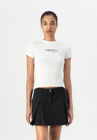 Haut court à manches courtes, blanc et à côtes, avec le logo "TOMMY JEANS New York" en bleu foncé, associé à une jupe mini plissée noire.