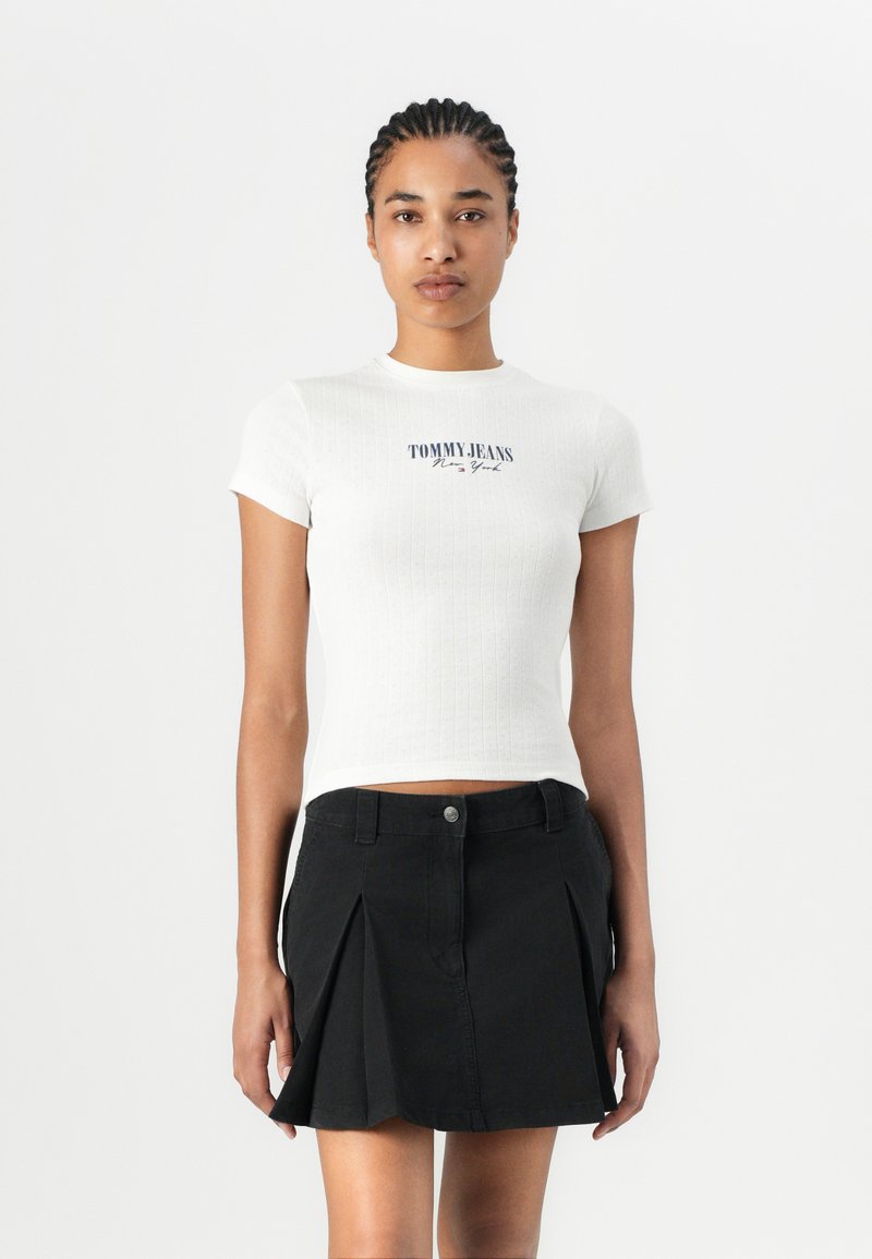 Haut court à manches courtes, blanc et à côtes, avec le logo "TOMMY JEANS New York" en bleu foncé, associé à une jupe mini plissée noire.