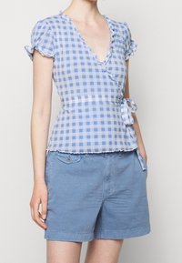 Top avvolgente a quadri blu con bordi arricciati, maniche corte a sbuffo e un nodo in vita. Abbinato a pantaloni corti di cotone azzurri.