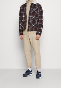 Veste à carreaux avec col en peau lainée, pull en maille beige, pantalon beige et baskets bleu marine. Présente des nuances variées de rouge, gris et blanc.