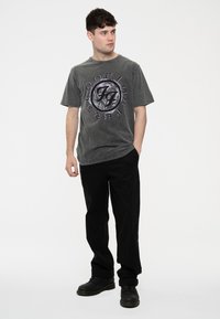 Gray cotton t-shirt with a circular black and purple logo print on the chest; paired with black pants and black boots.