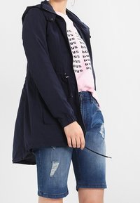 Granatowa kurtka z kapturem, z elastycznymi mankietami, troczkami w talii i kieszeniami z przodu, założona na różową koszulkę z grafiką, w połączeniu z postarzanymi niebieskimi szortami dżinsowymi.