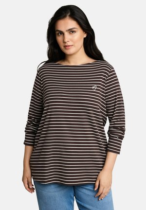 Frau mit langen dunklen Haaren, die ein braun-weiß gestreiftes Langarmshirt mit kleinem Herz-Design und blaue Jeans trägt, steht vor einem schlichten Hintergrund.
