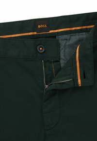 Pantaloni de culoare verde închis cu fermoar, accente de cusături portocalii, buton negru și o etichetă din țesătură cu "BOSS". Căptușeala interioară texturată este vizibilă.