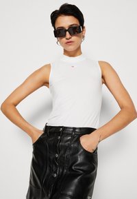 White sleeveless top with a high collar and red logo, paired with a black leather skirt featuring a front button closure and pockets.