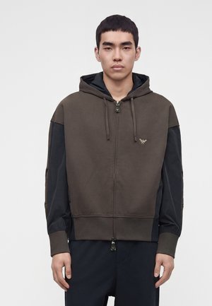 Jeune homme portant un sweat à capuche zippé bicolore marron et noir avec un petit logo aigle sur la poitrine gauche, debout devant un fond uni.