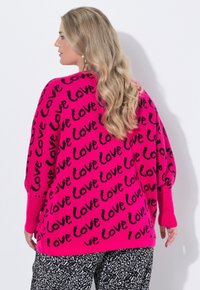 Světle růžový oversized svetr s opakujícím se černým vzorem textu "láska", žebrovanými manžetami a volným střihem. V kombinaci s černými kalhotami se vzorem.