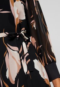 Robe portefeuille noire à motifs floraux, en tissu transparent avec de grandes fleurs roses et blanches, une ceinture à la taille et des poignets de manches élastiqués d'une couleur sombre unie.