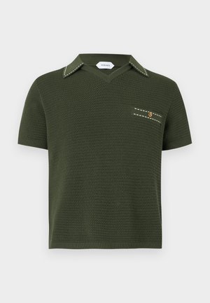 Mees, kes kannab rohelist lühikeste varrukatega kootud polosärki, musti mansettidega pükse ja musti paeltega saapaid, seisab käed taskutes tavalise tausta ees.