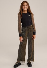 Svart långärmad topp med genomskinliga ärmar i metalltråd, matchad med guld- och svarta plisserade vida byxor. Svarta loafers med guldaccenter.