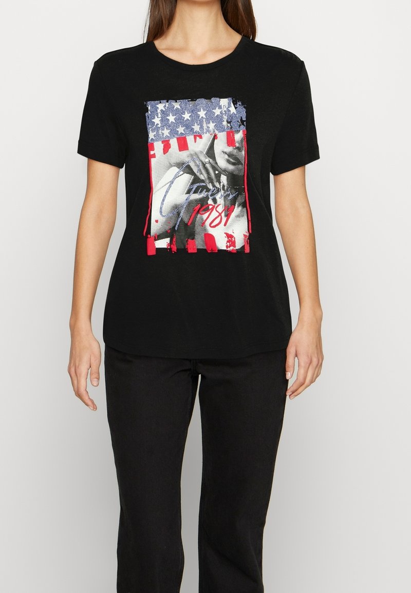 Femme portant un T-shirt noir avec une impression graphique du drapeau américain, un visage en niveaux de gris, et le texte "Guess 1981" en rouge et blanc.
