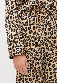 Satin pidžama set u leopard uzorku s bež pozadinom i crnim i smeškim mrljama. Uključuje dugme na manžetama i pojas s elastikom.