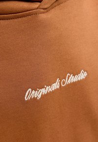 Rjava tkanina z gladko teksturo in povišeno šivanje. "Originals Studio" je izvezena v belem kurvikastem tisku.