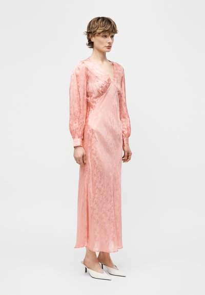 Γυναίκα που φοράει ένα μακρύ ροζ σατέν φόρεμα με φουσκωτά μακριά μανίκια και λευκές μυτερές mules με τακούνι, στέκεται μπροστά σε λευκό, απλό φόντο.