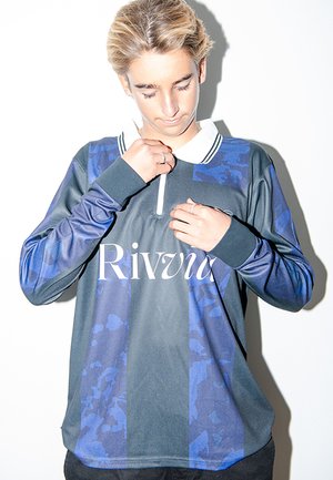 Νεαρός με κοντά ξανθά μαλλιά που προσαρμόζει το γιακά σε μπλε και μαύρη ριγέ αθλητική μπλούζα με το λογότυπο "Rivii".