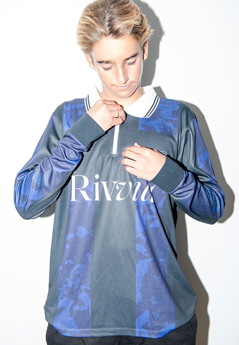 Νεαρός με κοντά ξανθά μαλλιά που προσαρμόζει το γιακά σε μπλε και μαύρη ριγέ αθλητική μπλούζα με το λογότυπο "Rivii".