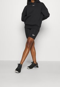 Haut en molleton oversize noir avec un logo, associé à un short noir arborant un logo. Chaussures de sport noires avec des accents gris, posant devant un fond neutre.