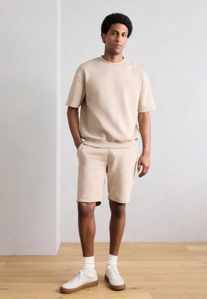 Rövid göndör hajú férfi, bézs rövid ujjú pulóvert és rövidnadrágot visel, fehér zoknival és fehér tornacipővel, fapadlón áll.