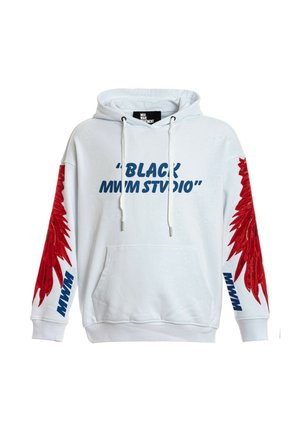 Joven con sudadera blanca con capucha y diseños de alas rojas en las mangas y texto azul, pantalones cortos rojos con cadena, calcetines blancos y zapatos beige.