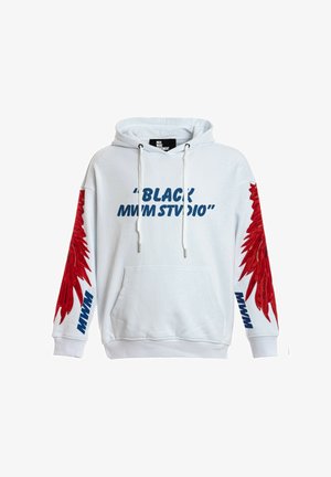 Joven con sudadera blanca con capucha y diseños de alas rojas en las mangas y texto azul, pantalones cortos rojos con cadena, calcetines blancos y zapatos beige.