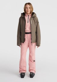 Dames skikleding met een bruin, geprofileerd jack, roze geïsoleerde broek en zwarte skischoenen met verstelbare riemen.