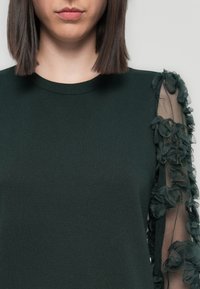 Green sweatshirt with a round neckline and long sleeves. One sleeve features a sheer fabric decorated with layered floral appliqués.
