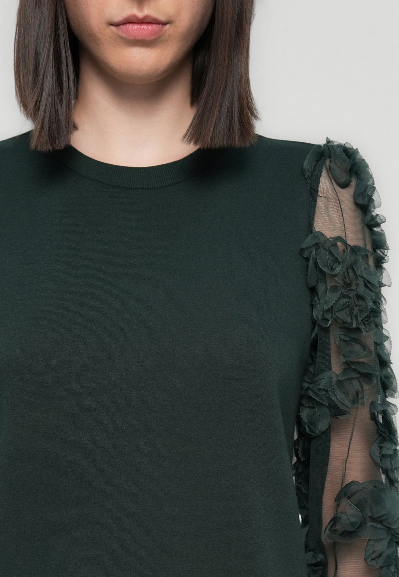 Grüner Pullover mit rundem Ausschnitt und langen Ärmeln. Ein Ärmel hat ein transparentes Gewebe, das mit gestuften Blumenapplikationen verziert ist.