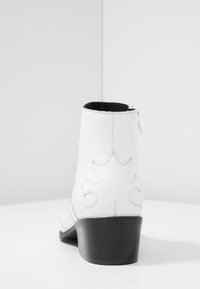 Botín de cuero blanco hasta el tobillo con suela negra y sutil costura decorativa, mostrado desde la parte posterior sobre un fondo blanco liso.