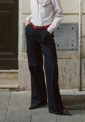 Dunkelblaue, hoch taillierte Jeans mit Falten, kombiniert mit einem weißen Hemd mit Knöpfen und Rüschenkragen sowie einem roten Gürtel, ergänzt durch spitze Schuhe.