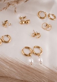 Cerceii în tonuri aurii prezintă diferite modele, inclusiv cercuri cu accente de perle, cercei cu șurub și piese cu forme jucăușe, așezate pe un fundal moale de țesătură.