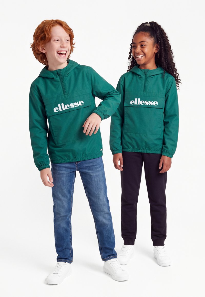 Zwei lächelnde Kinder tragen passende grüne ellesse Kapuzenjacken, eines in Jeans und das andere in schwarzen Hosen und weißen Schuhen.