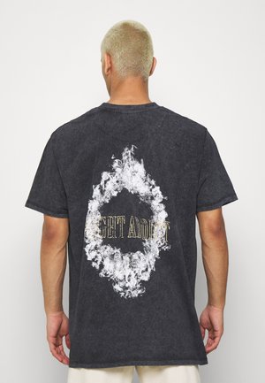 Osoba o krótkich blond włosach, ubrana w czarny t-shirt z białym, dymnym owalem i częściowo widocznym żółtym napisem na plecach.