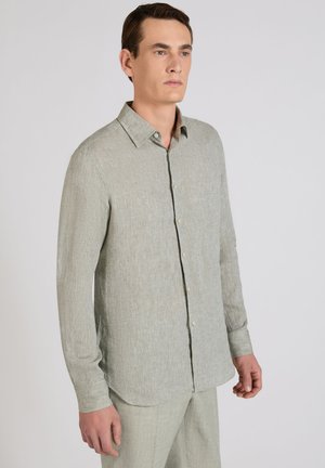 Homme portant une chemise à boutons à manches longues gris clair et un pantalon assorti, debout devant un fond clair uni.