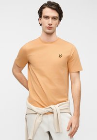 Κοντομάνικο, ροδακινί T-shirt από απαλό βαμβάκι, με μικρό μαύρο λογότυπο πουλιού στο στήθος, χαλαρή εφαρμογή και στρογγυλή λαιμόκοψη.