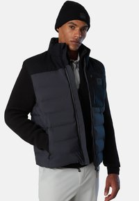 Steppweste in Dunkelgrau und Schwarz, mit gerippten Ärmeln, Reißverschlusstaschen und hohem Kragen, getragen über einem weißen Hemd. Schwarze Mütze auf dem Modell.