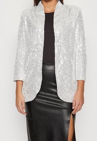 Kvinna iklädd en silverfärgad paljettblazer över en svart topp och en svart läderskjort med hög slits, står med avslappnade armar.