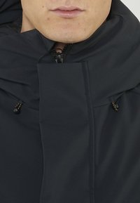 Giacca a vento navy con cappuccio, dotata di chiusura a zip, coulisse regolabili e una texture liscia e opaca. Dettagli cucito lungo la parte anteriore.