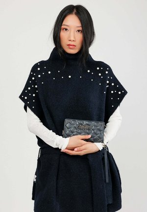 Marineblauer Wollponcho mit hohem Kragen, verziert mit weißen Perlen an den Schultern, kombiniert mit einer schwarzen, gesteppten Clutch mit goldenen Akzenten.