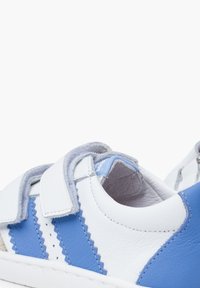 Scarpe in pelle bianche e azzurre chiare con un cinturino in velcro, caratterizzate da motivi a zigzag sui lati e una finitura di texture.