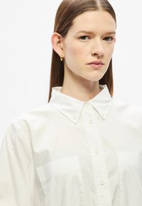 Fehér pamut ing hosszú ujjakkal, gombos gallérral és két mellzsebbel. Sima textúra és klasszikus szabás. Finom fényesség.