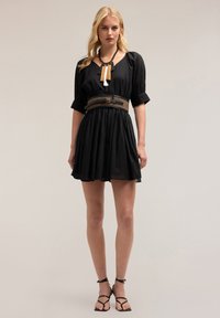 Schwarzes kurzes Kleid mit weit ausgestellt Rock, V-Ausschnitt und Puffärmeln. Akzentuiert mit einem geflochtenen Gürtel und einer geschichteten Kette mit Quasten.