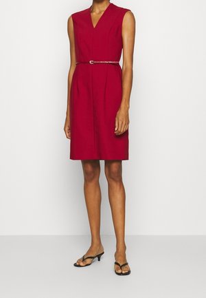 Ärmelloses rotes knielanges Kleid mit V-Ausschnitt und dünnem passendem Gürtel, getragen mit schwarzen Sandalen mit niedrigem Absatz und offenem Zehenbereich.