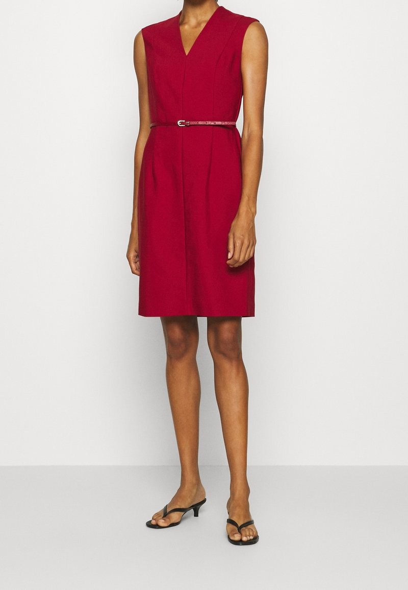 Robe rouge sans manches longueur genou avec col en V et fine ceinture assortie, portée avec des sandales noires à talons bas et bout ouvert.