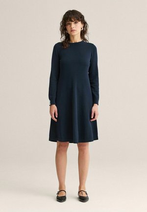 Kvinde står iført en knælang, langærmet navyblå kjole med sorte Mary Jane-sko foran en ensfarvet beige baggrund.