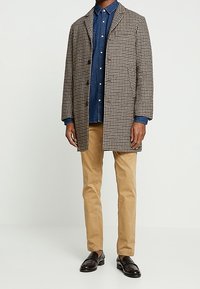 Hahnentritt-geprägter Überziehmantel in Braun und Creme, über einem Jeanshemd getragen. Kombiniert mit beigen Chinos und schwarzen Slippern.