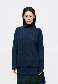 Pulover din tricot cu guler înalt, de culoare bleumarin, cu model cable-knit, având tivuri și mansete ribonate. Prezintă un design texturat și un detaliu mic cu logo pe piept.