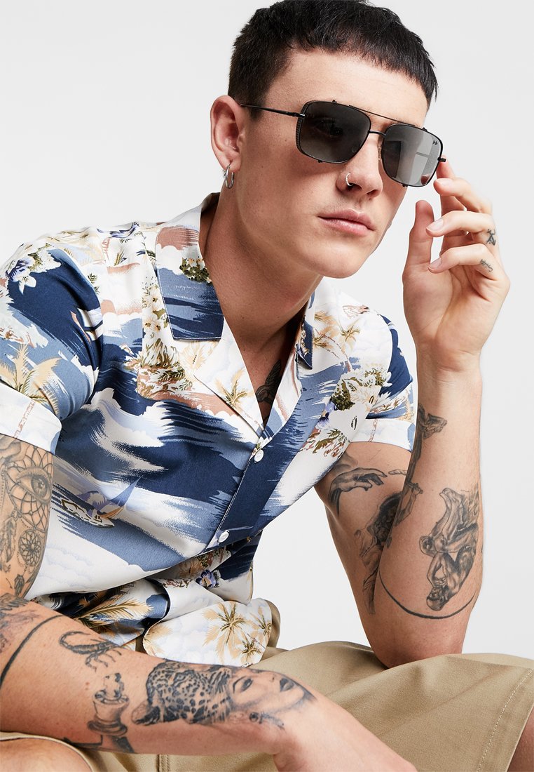 Korte mouw shirt met een bloemen- en abstract blauw patroon, voorzien van een kraag en knoopsluiting. Vergezeld van donkere zonnebrillen.