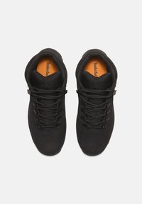 Black footwear with a textured upper, featuring round laces, padded collar, and contrasting orange insole with "Timberland" branding.